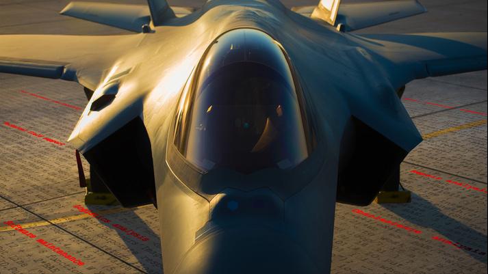 An F-35 Lightning II fighter aircraft sits on the ground. The ground is overlaid with software code.