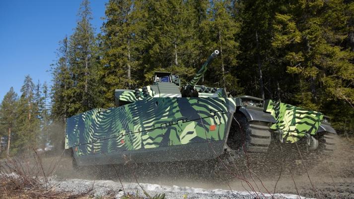 The combat-proven, camouflaged CV90 MkIV is in motion on a gravel track in front of a green forest. 